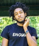 Young man with curly hair and a beard wearing a dark T-shirt with the word 'ENOUGH' printed on it, standing outdoors with trees in the background.