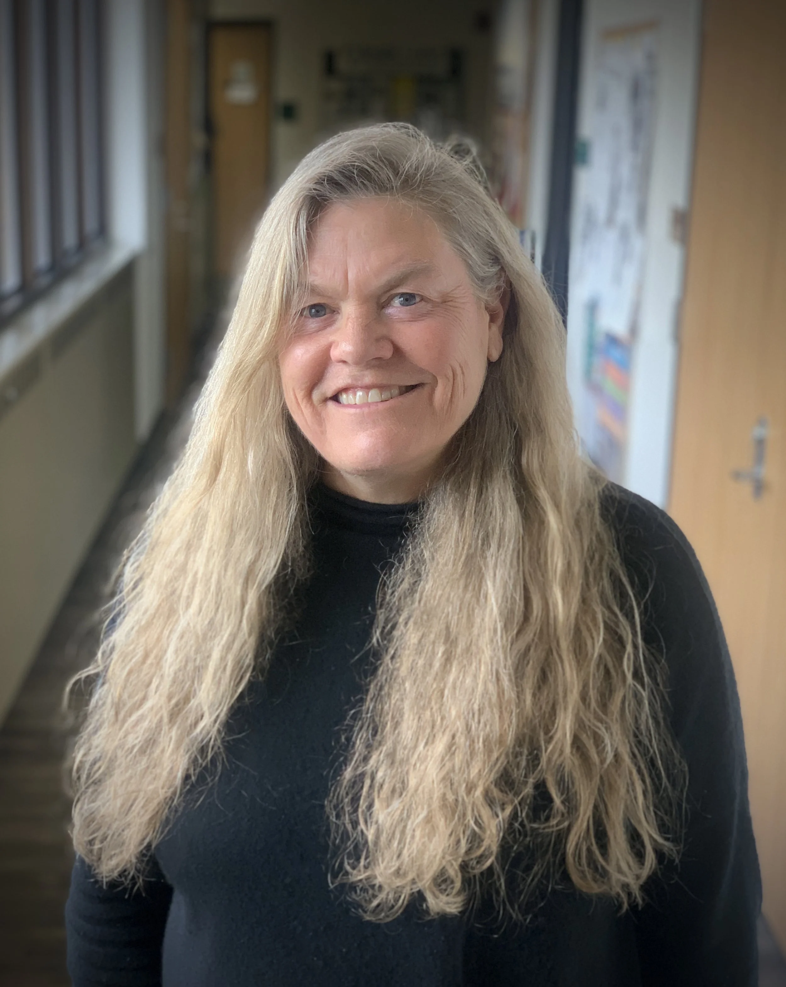 A smiling woman with long blonde hair wearing a black top inside a corridor with windows on one side and bulletin boards on the other.