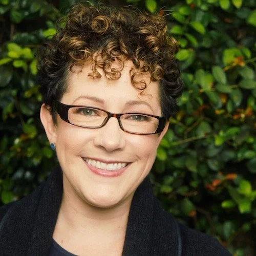 A woman with short curly hair, wearing glasses and a black jacket, smiling outdoors with green foliage in the background.