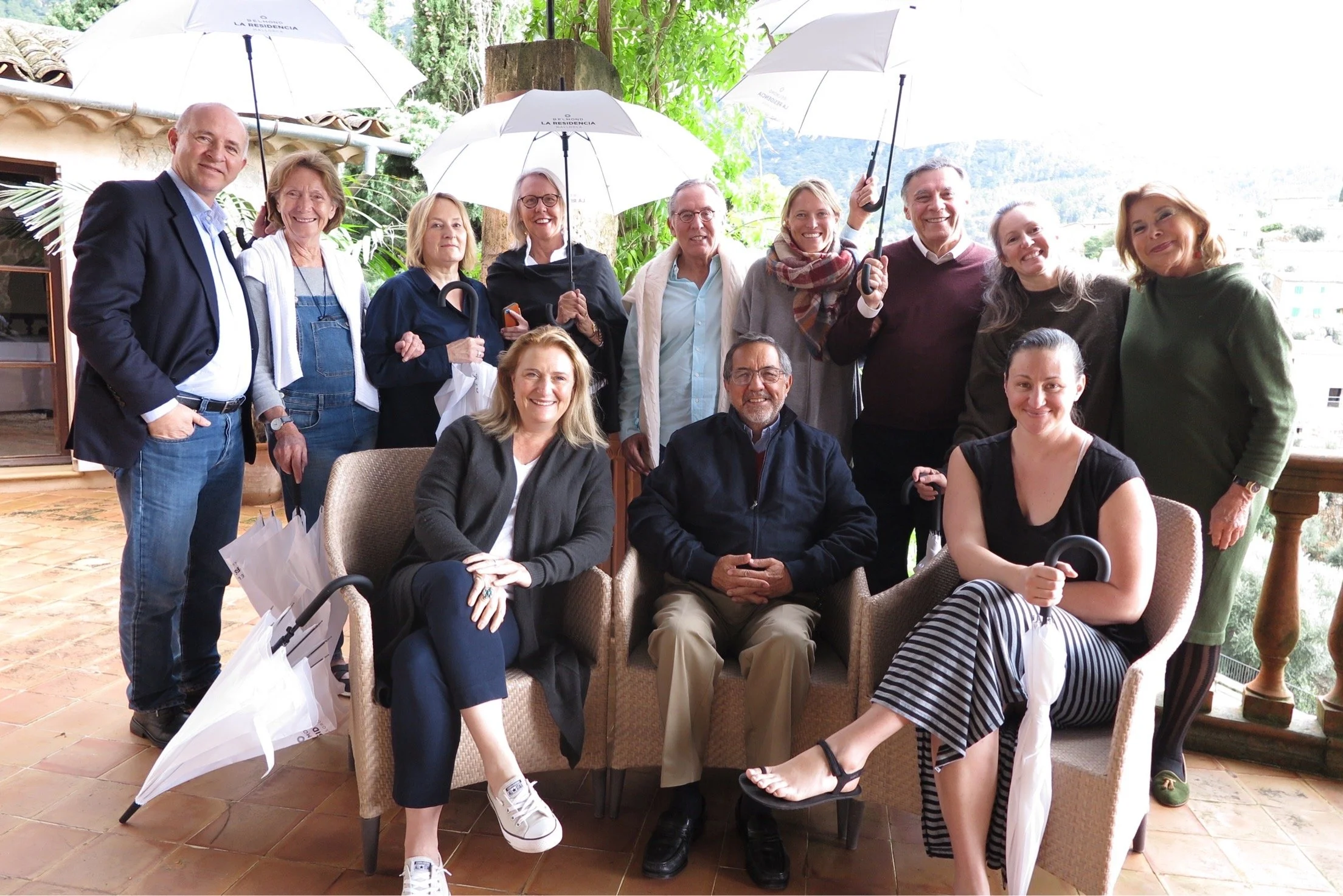 A group of 13 people gathered outdoors on a patio, some holding umbrellas, smiling at the camera.