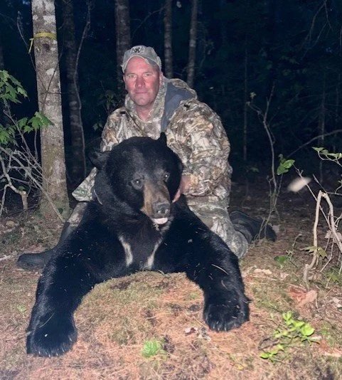 A man in camouflage clothing kneeling behind a large black bear he appears to have hunted, in a forested area at night.