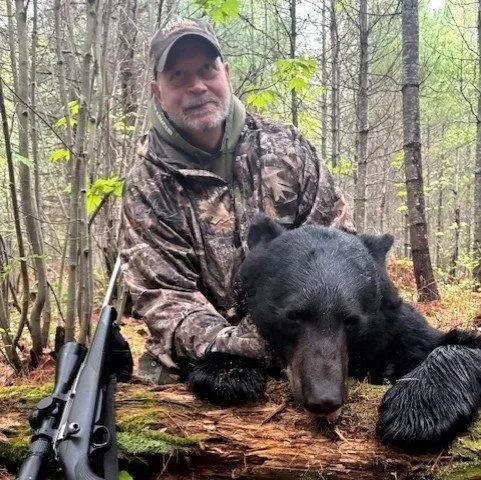 Man in camouflage clothing holding a large black bear in a forested area.