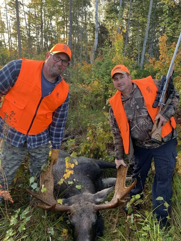 Two men dressed in camouflage and orange hunting gear posing with a large dead moose in a wooded area.