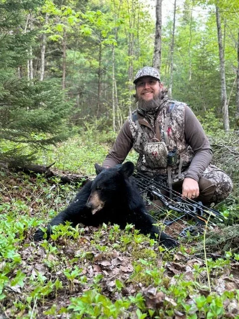 A man in camouflage hunting gear kneeling next to a black bear he has hunted, in a green forest with trees and ground cover.