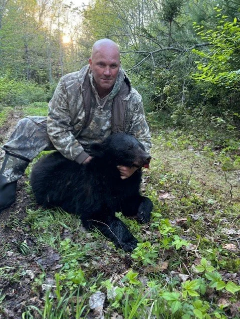 A man in camouflage clothing kneels on the ground in a forest, holding a black bear cub.