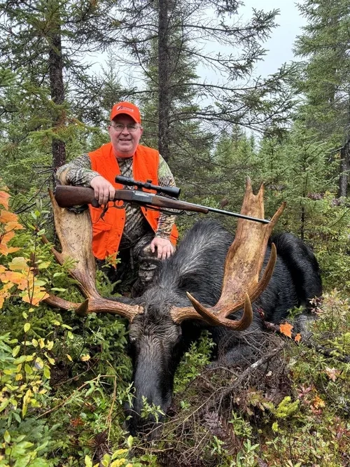 A man in camouflage clothing and an orange hunting vest and hat kneels next to a large moose with impressive antlers in a forested area, holding a rifle.
