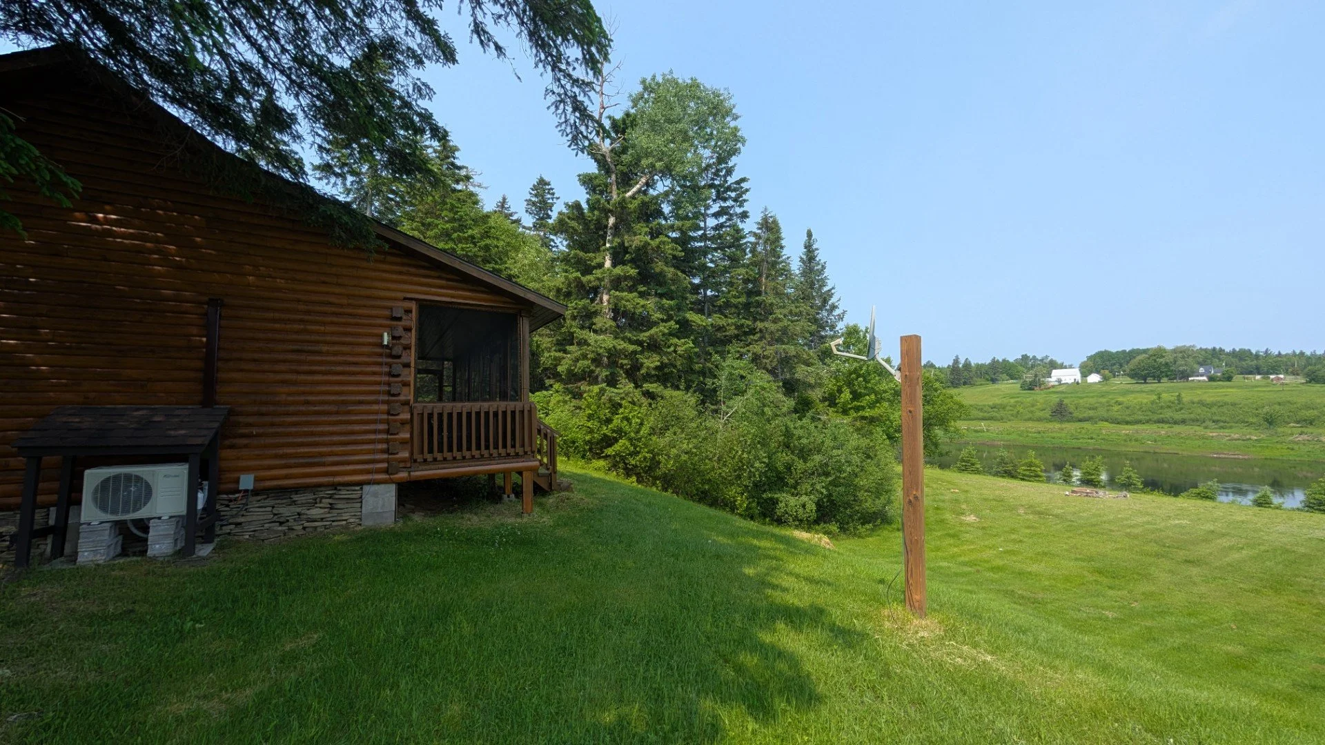 Photograph of a wooden cabin with a small porch on a lush green grassy backyard beside a pond, surrounded by trees and hills, under a blue sky.