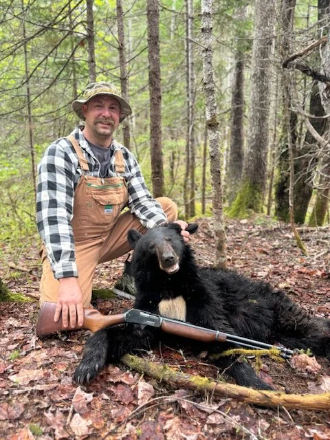 A man in outdoor gear kneeling beside a black bear lying on the forest ground, with a rifle resting nearby.