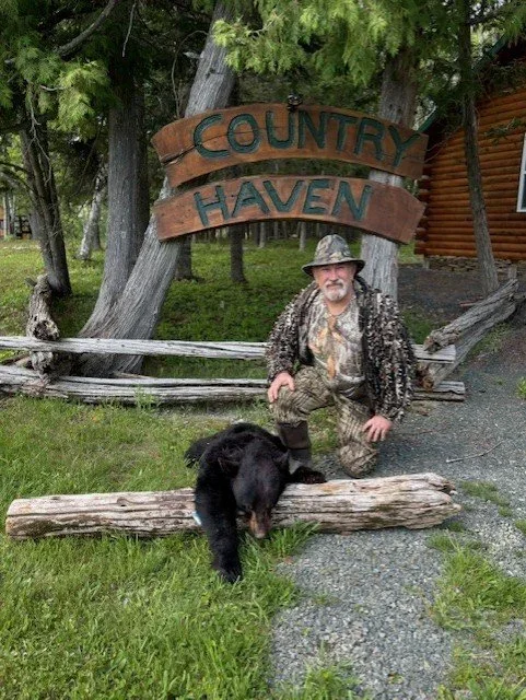 A man dressed in camouflage and a camouflage hat kneels next to a black and brown dog on a log in front of a wooden sign that reads "Country Haven". The scene is outdoors with trees and a log fence in the background.