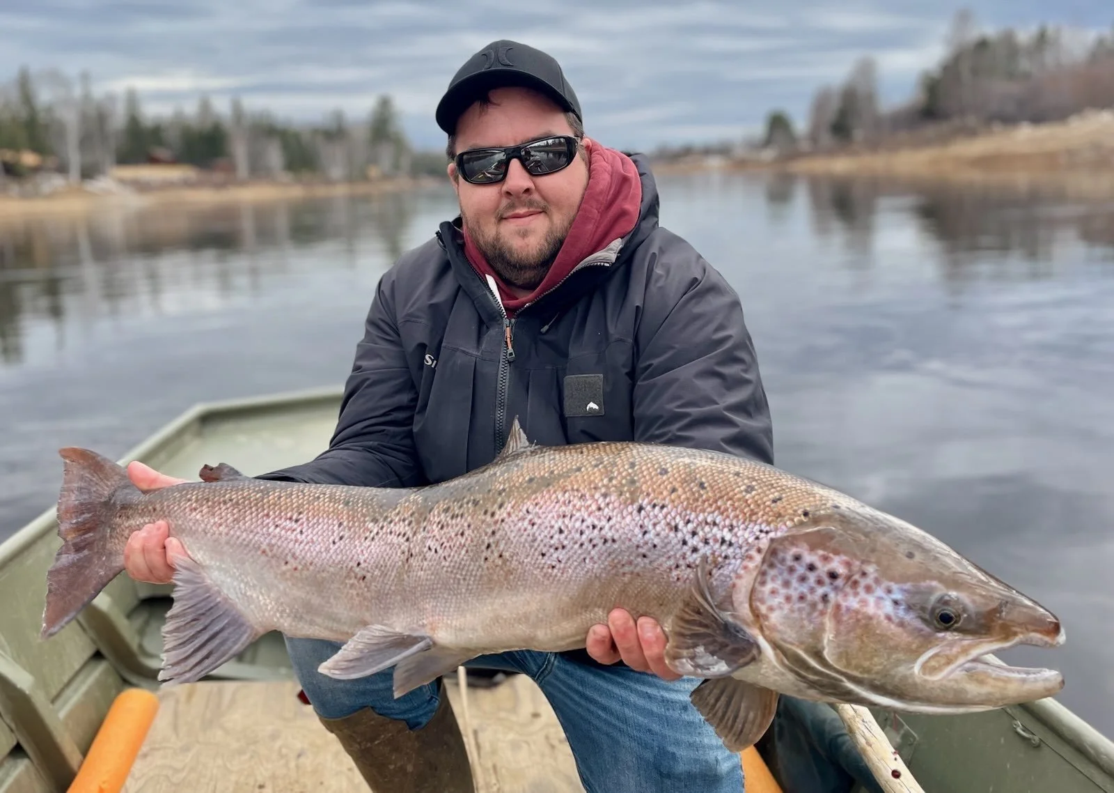 A man wearing sunglasses, a black hat, and a dark jacket holding a large fish on a boat with a calm river and trees in the background.
