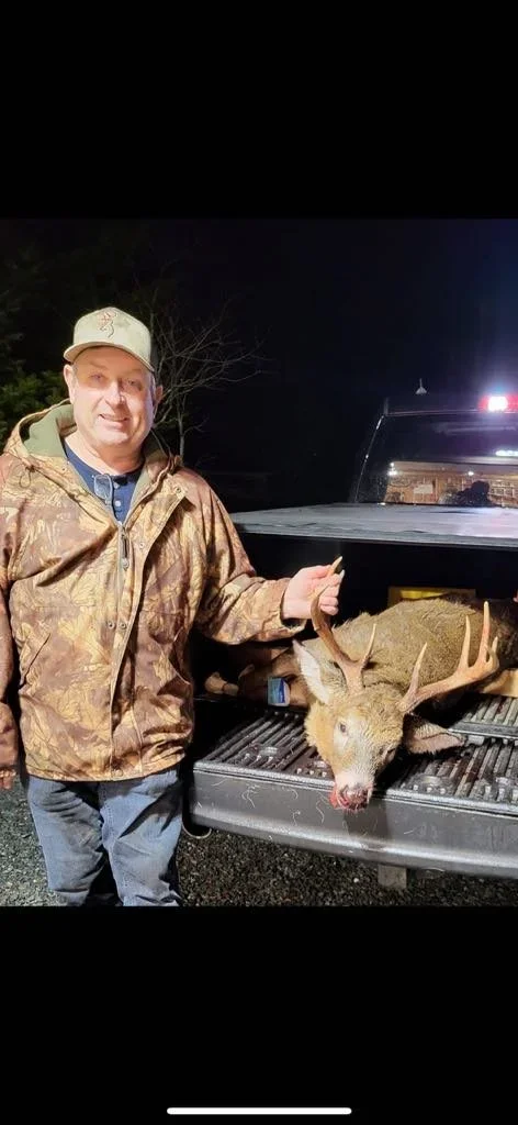 A man in camouflage jacket and beige cap standing next to a pickup truck with a large deer with antlers lying in the truck bed, taken at night.