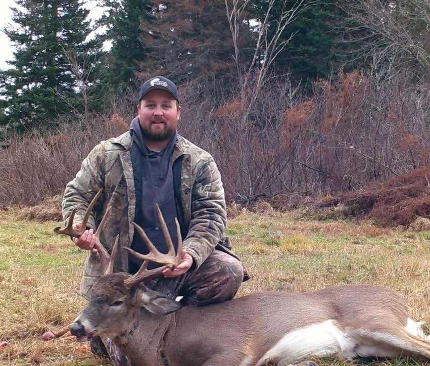 A man in camouflage clothing holds the antlers of a large, freshly hunted deer lying on the ground in a field with trees in the background.