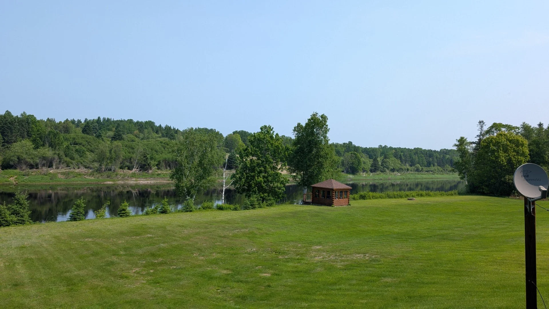 A grassy lawn next to a river with trees in the background. A small wooden gazebo is near the water, and a satellite dish is on the right side of the image.