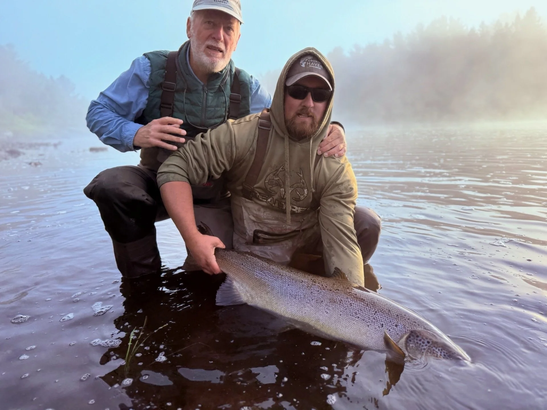 Two men fishing in a river, one holding a large fish, with foggy trees in the background.