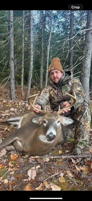 Man in camouflage clothing holding the antlers of a large deer with a forest in the background.