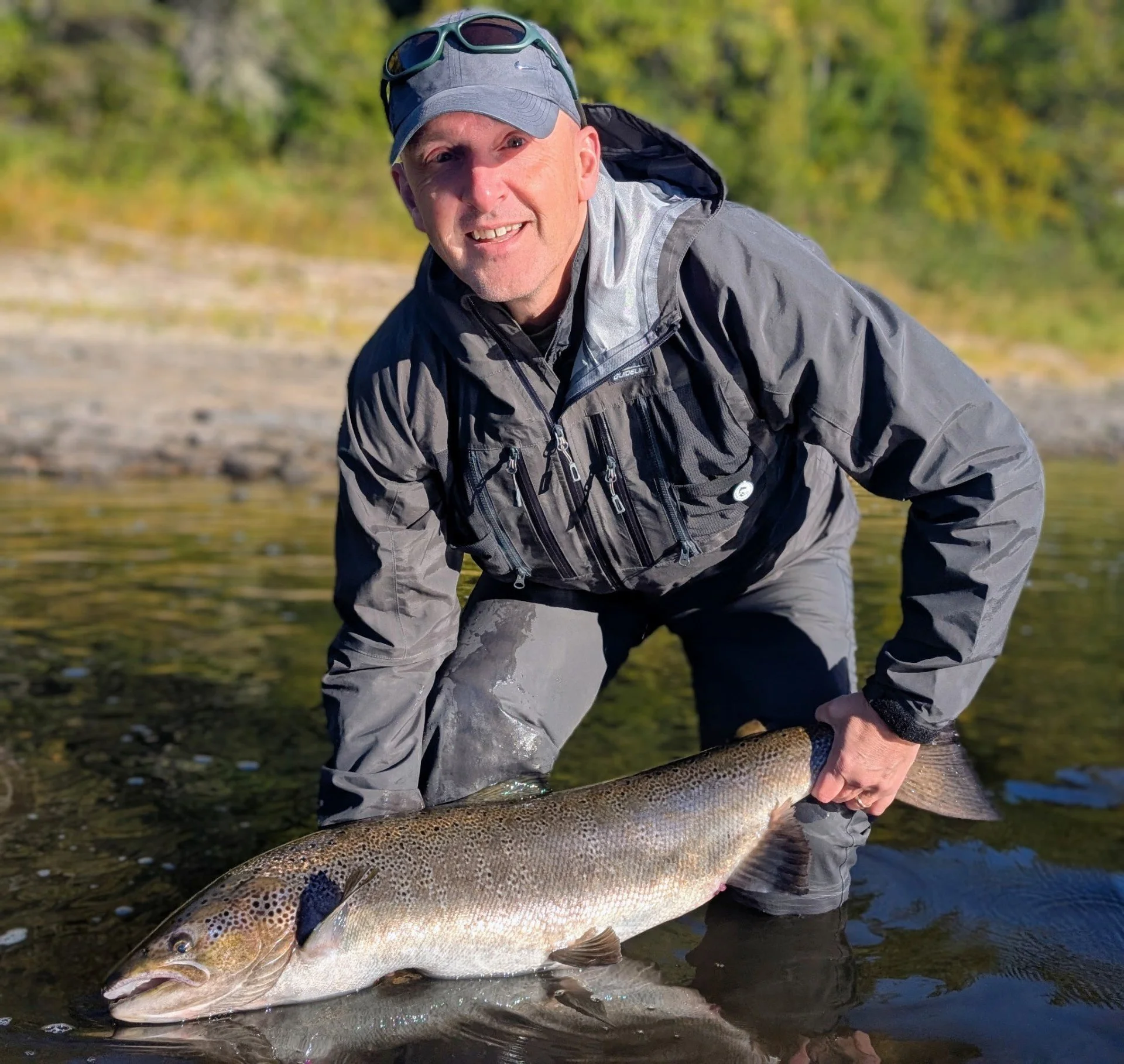 A man smiling, dressed in outdoor gear, holding a large fish near a riverbank.