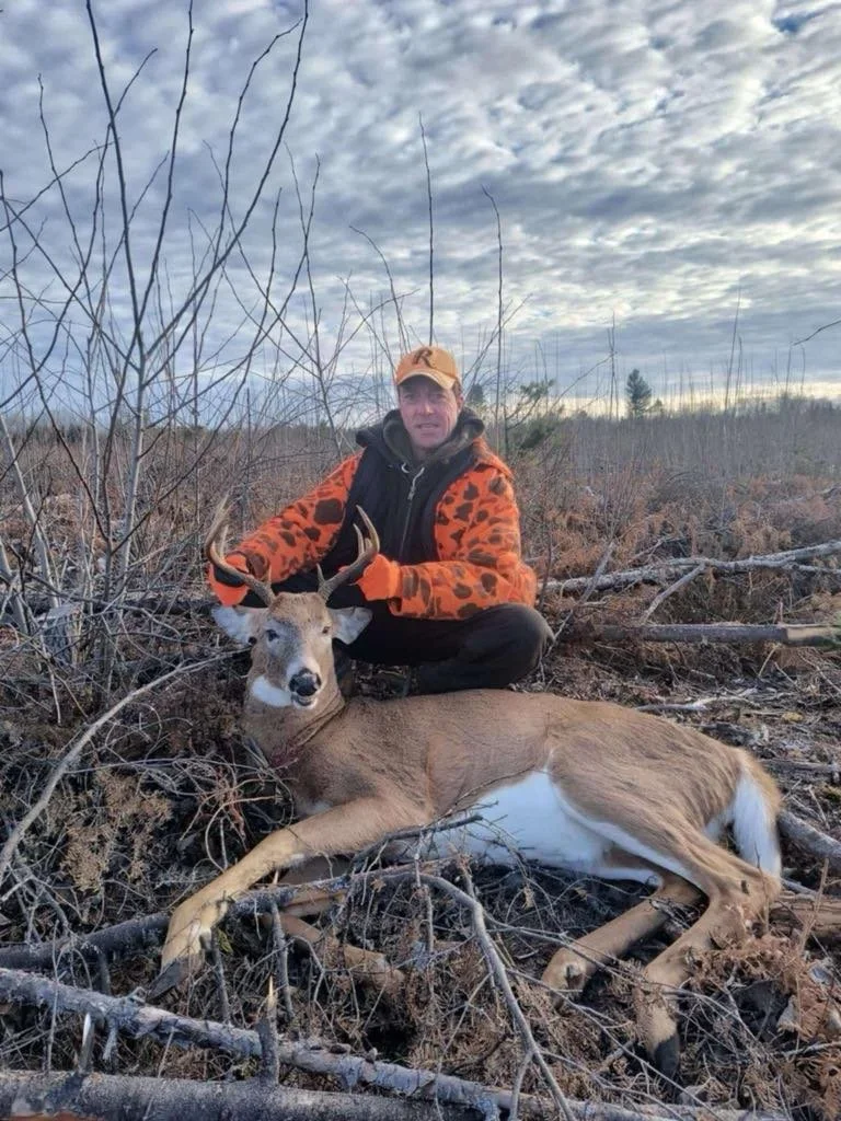 A man crouching behind a freshly hunted deer in a wooded area with leafless branches and a cloudy sky overhead.