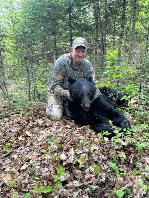 A man in camouflage clothing kneels in a forest, smiling, with a dead black bear lying on the ground beside him.