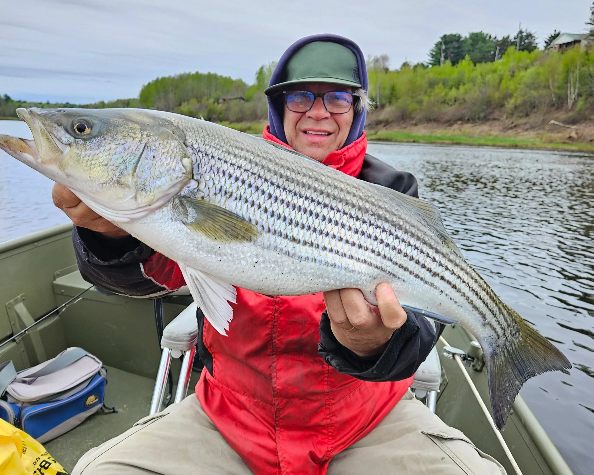 Man in a boat holding a large fish on a river, wearing glasses, a hat, and a red jacket.