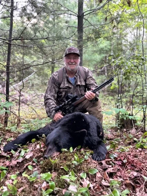 A man in camouflage clothing kneels in a forested area with a large black bear lying on the ground in front of him. He is holding a rifle and smiling.