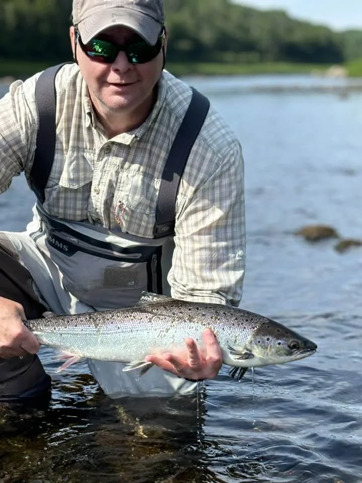 Man wearing sunglasses, hat, and fishing gear in a river holding a large fish.