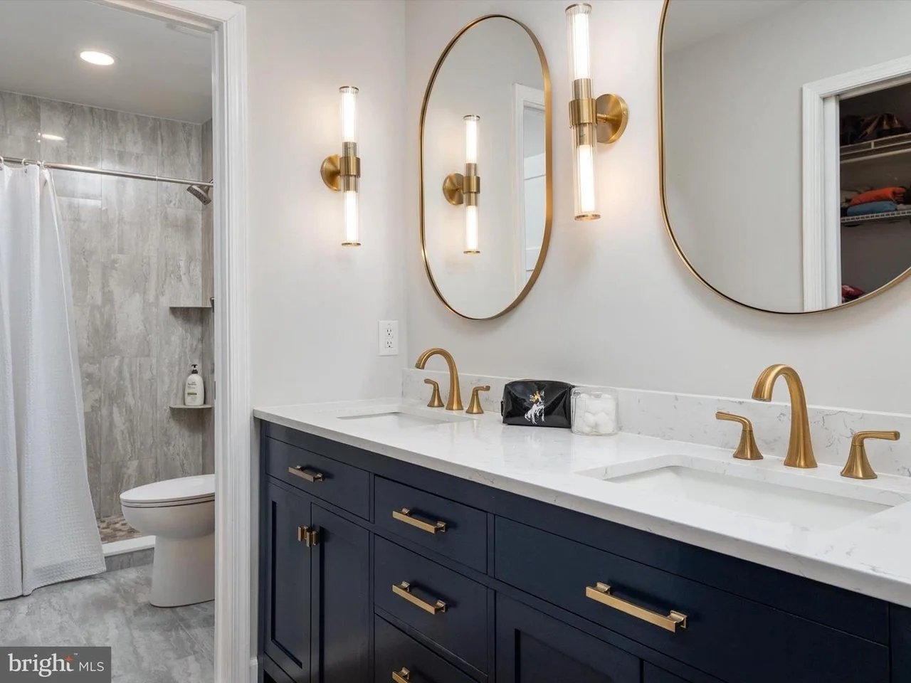Modern bathroom with navy vanity, white marble countertop, and double sinks with gold fixtures. Two oval mirrors and wall-mounted gold lights above the sinks. Visible walk-in shower with gray tiles and white shower curtain in the background.