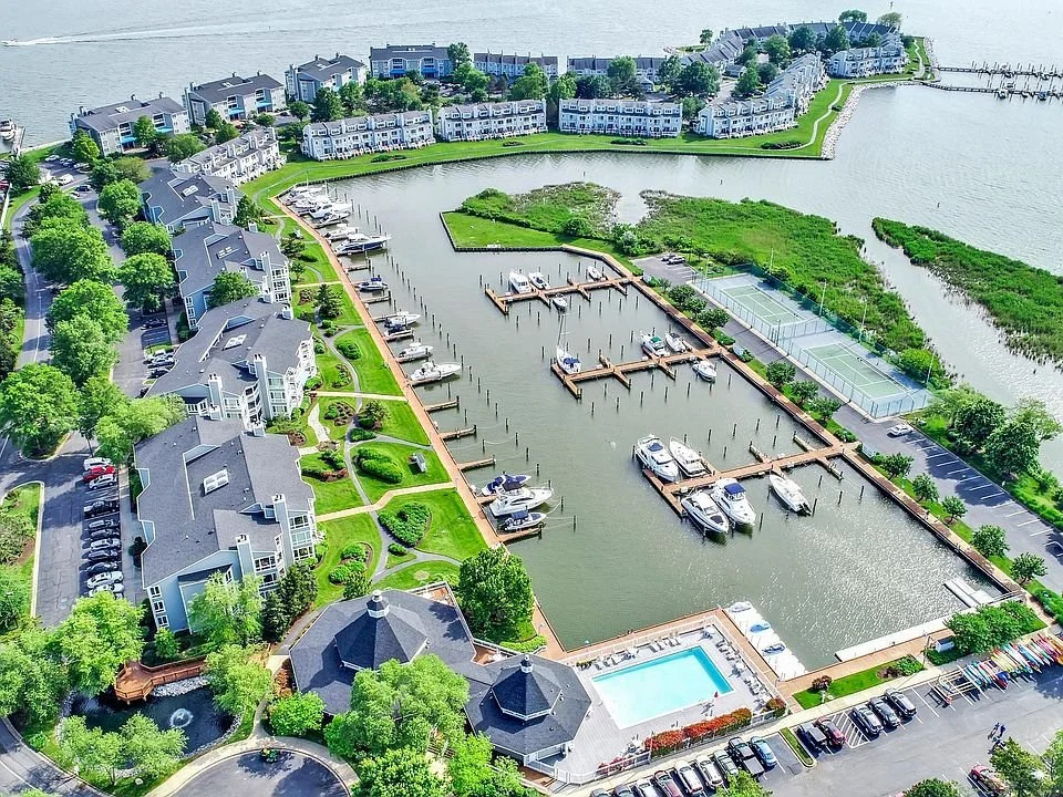 Aerial view of a waterfront residential complex with multiple townhouses, a marina with boats, tennis courts, a swimming pool, parking lots, and green landscaping along a water body.