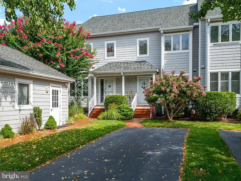 Front view of a gray townhouse with a small porch, white railing, and steps leading to the door, surrounded by green bushes and trees, with a paved driveway in front and a small separate building on the left. There are pink flowering trees on both si