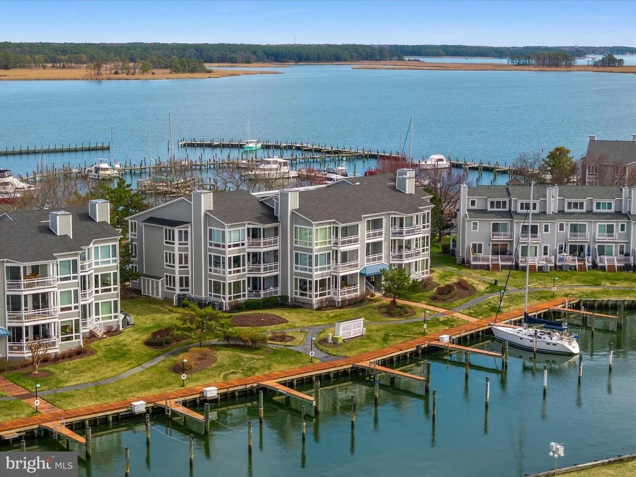 Aerial view of a lakeside residential area with boats docked at piers, surrounded by grass, trees, and multi-story apartment buildings.