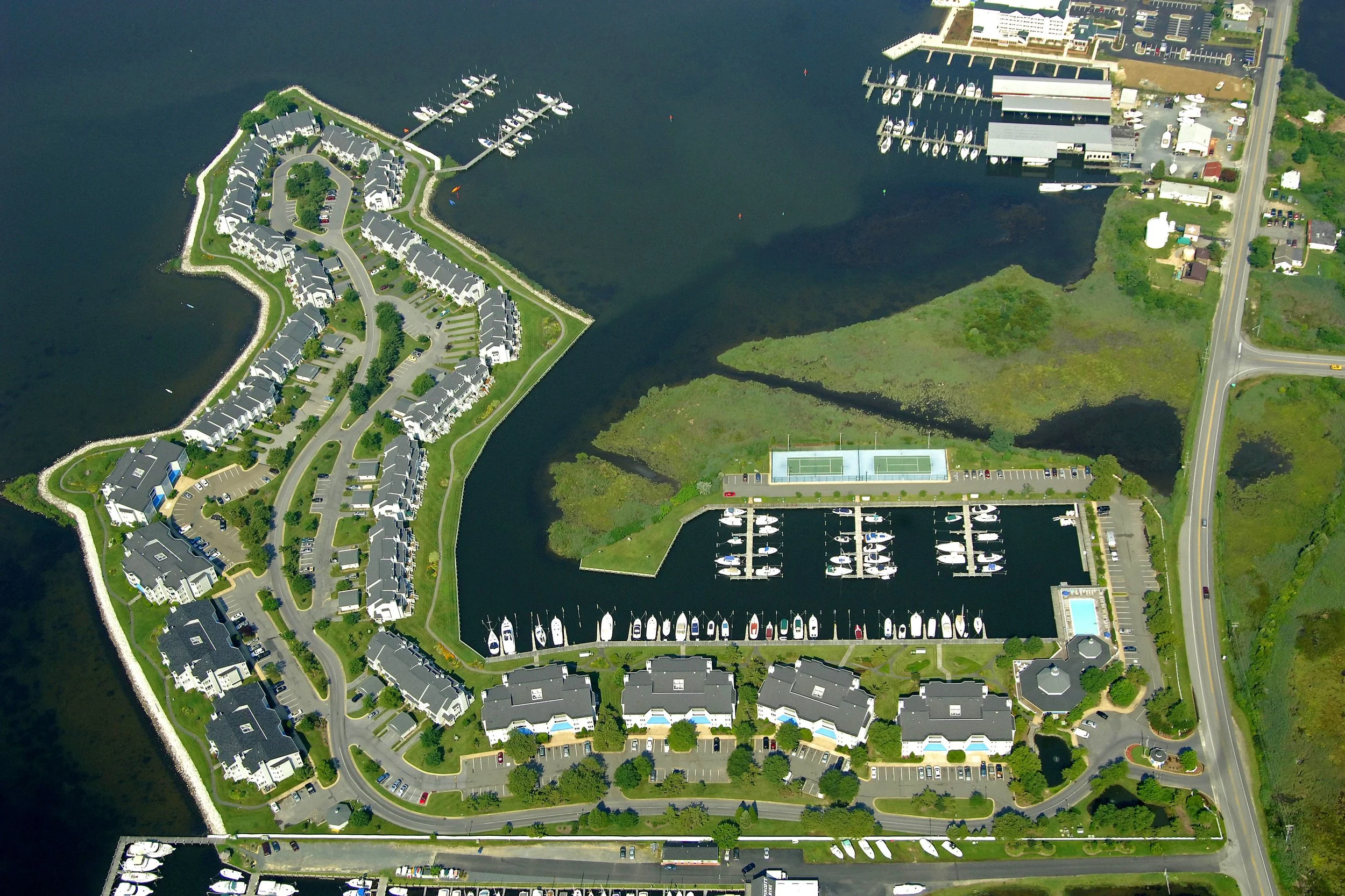 Aerial view of a marina with boats docked, residential apartments, tennis courts, and parking lots by the water, with surrounding roads and green areas.