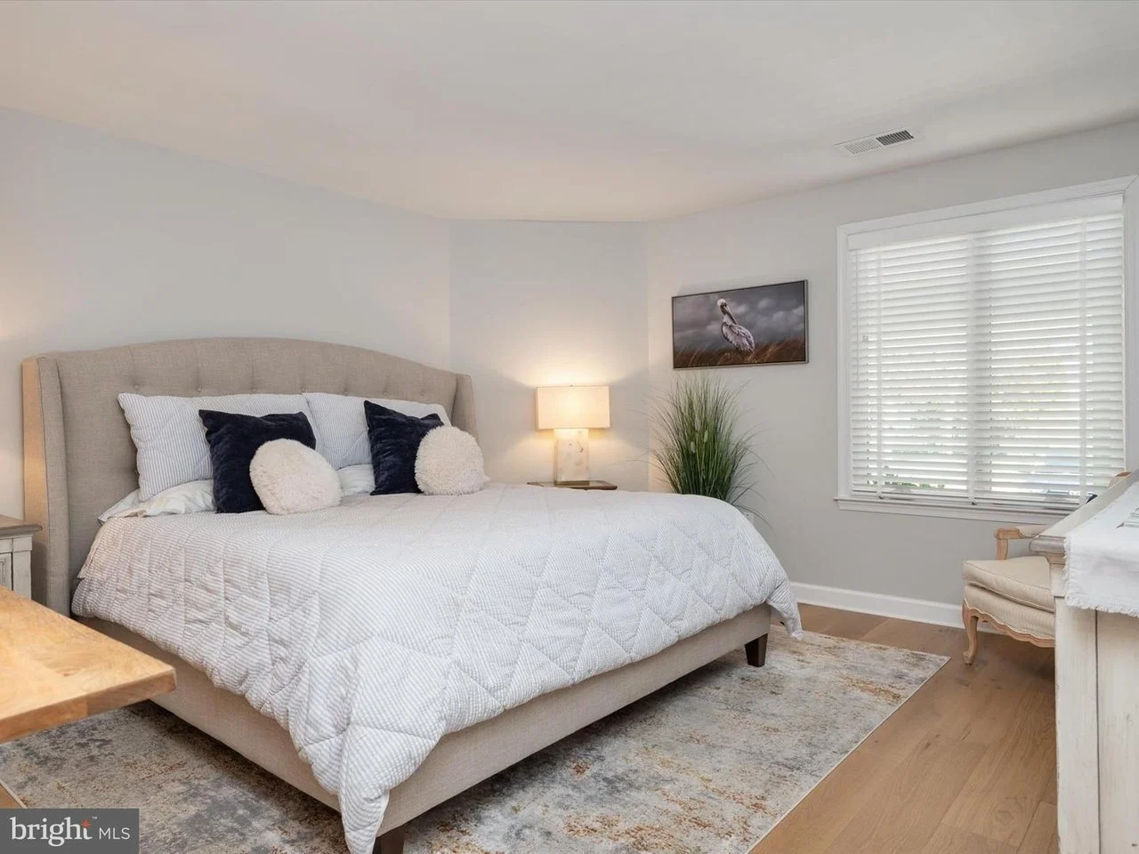 A bedroom with a beige upholstered bed, white bedding, decorative pillows, a nightstand with a lamp, a window with blinds, a wall photo of an egret, a green plant, and a chest of drawers.