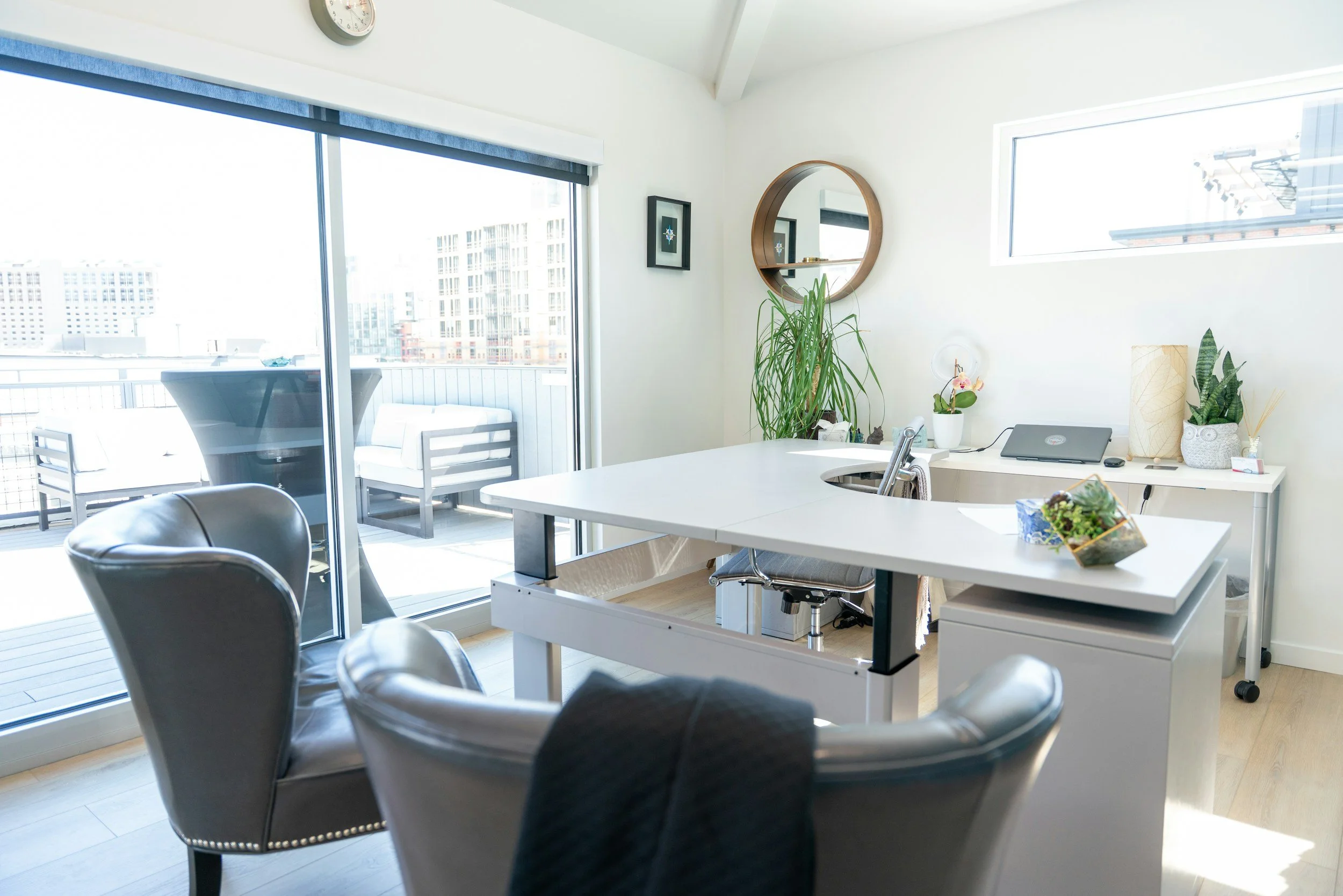 A bright, modern workspace with a white desk, off-center from the image, with a laptop, plants, and decor. There are black chairs in the foreground, a sliding glass door leading to a balcony with outdoor seating, and city buildings visible outside. A round mirror and small framed artwork are on the wall.