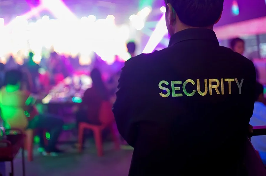 Security guard in black jacket with reflective 'SECURITY' lettering overseeing a crowded event with colorful lighting.