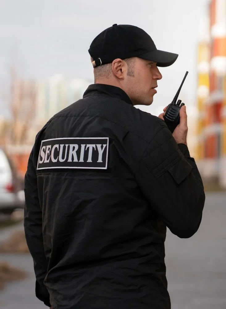 Security guard in black uniform and cap holding a walkie-talkie, standing outdoors near building or parking lot.