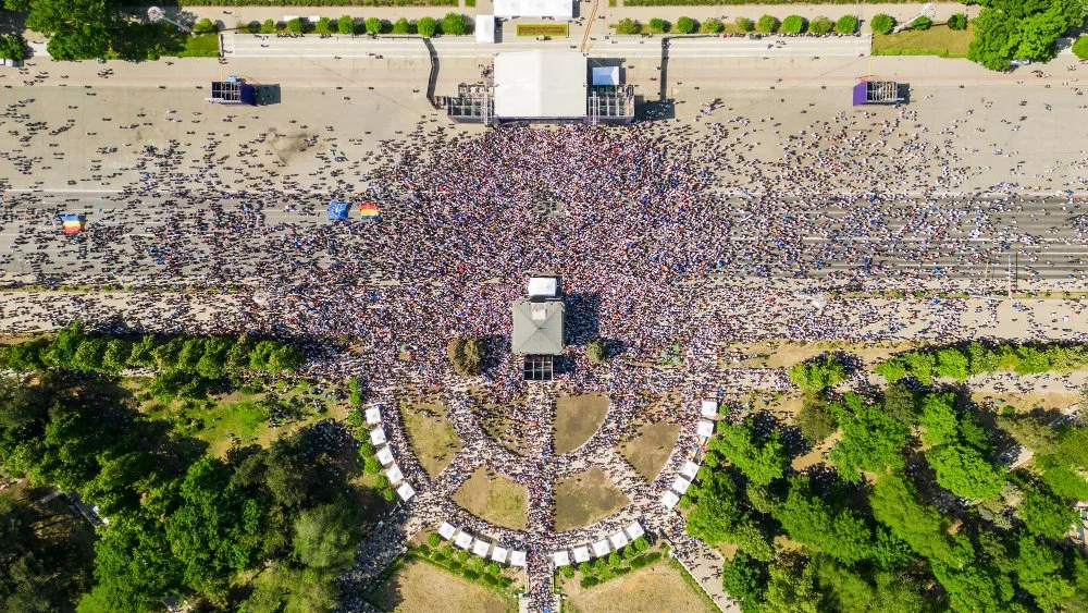 Aerial view of a large crowd gathered in a park area, with some tents and a building, surrounded by green trees and a street with more people.
