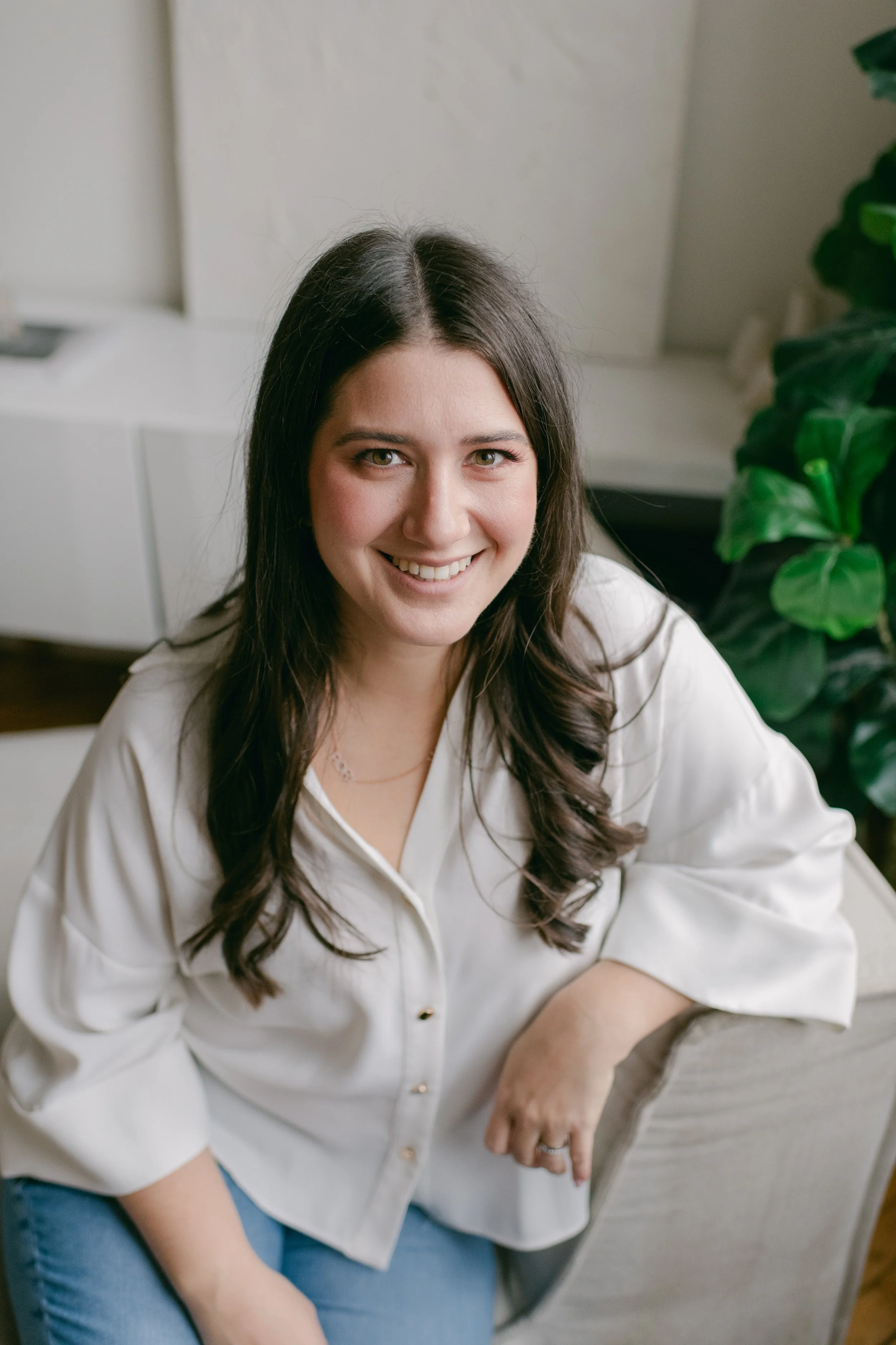 A woman with long dark hair sitting on a light-colored chair, smiling at the camera, with green plants in the background.