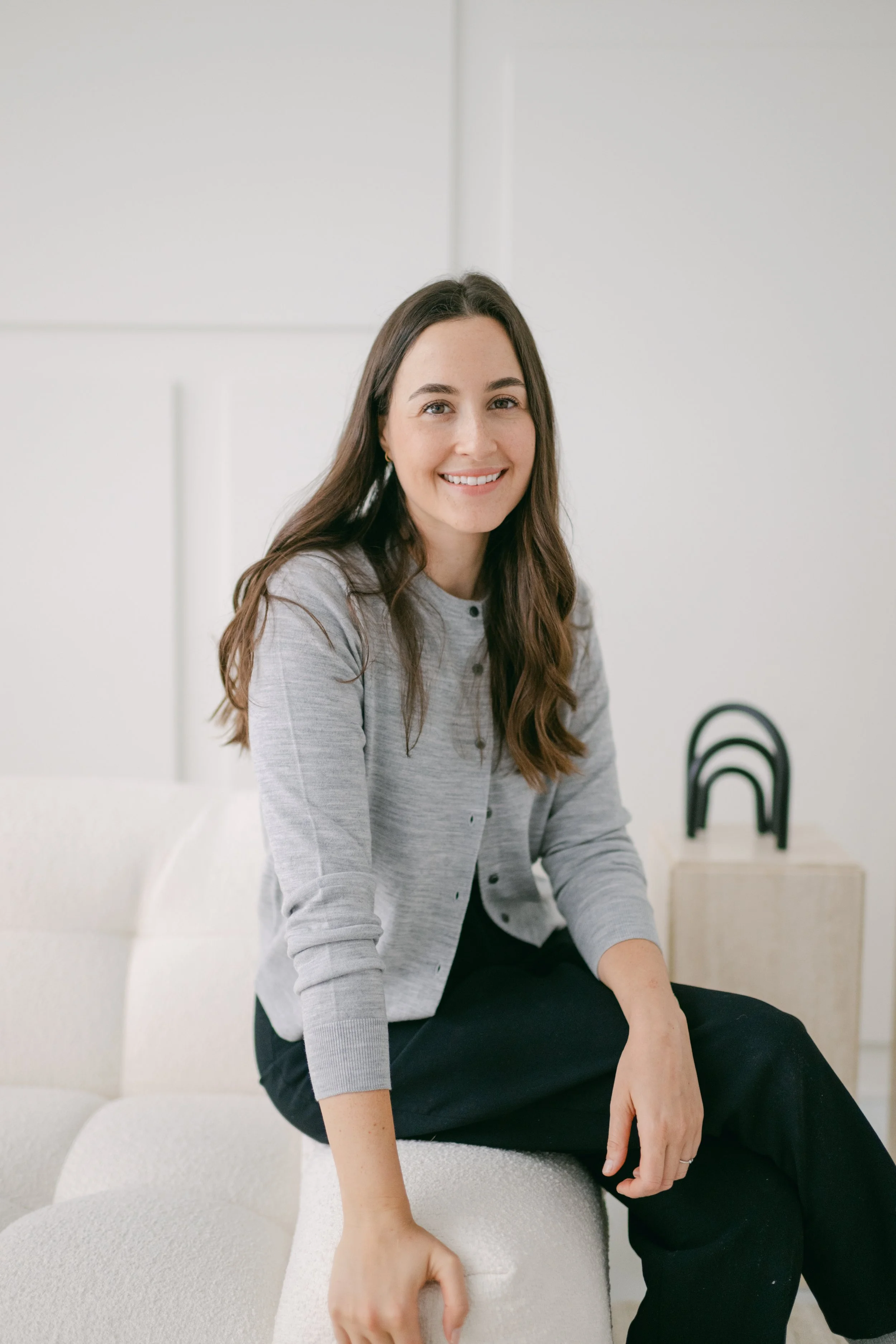 A young woman with long brown hair, wearing a gray cardigan and black pants, sitting on a white textured couch, smiling at the camera in a bright, minimalist room.