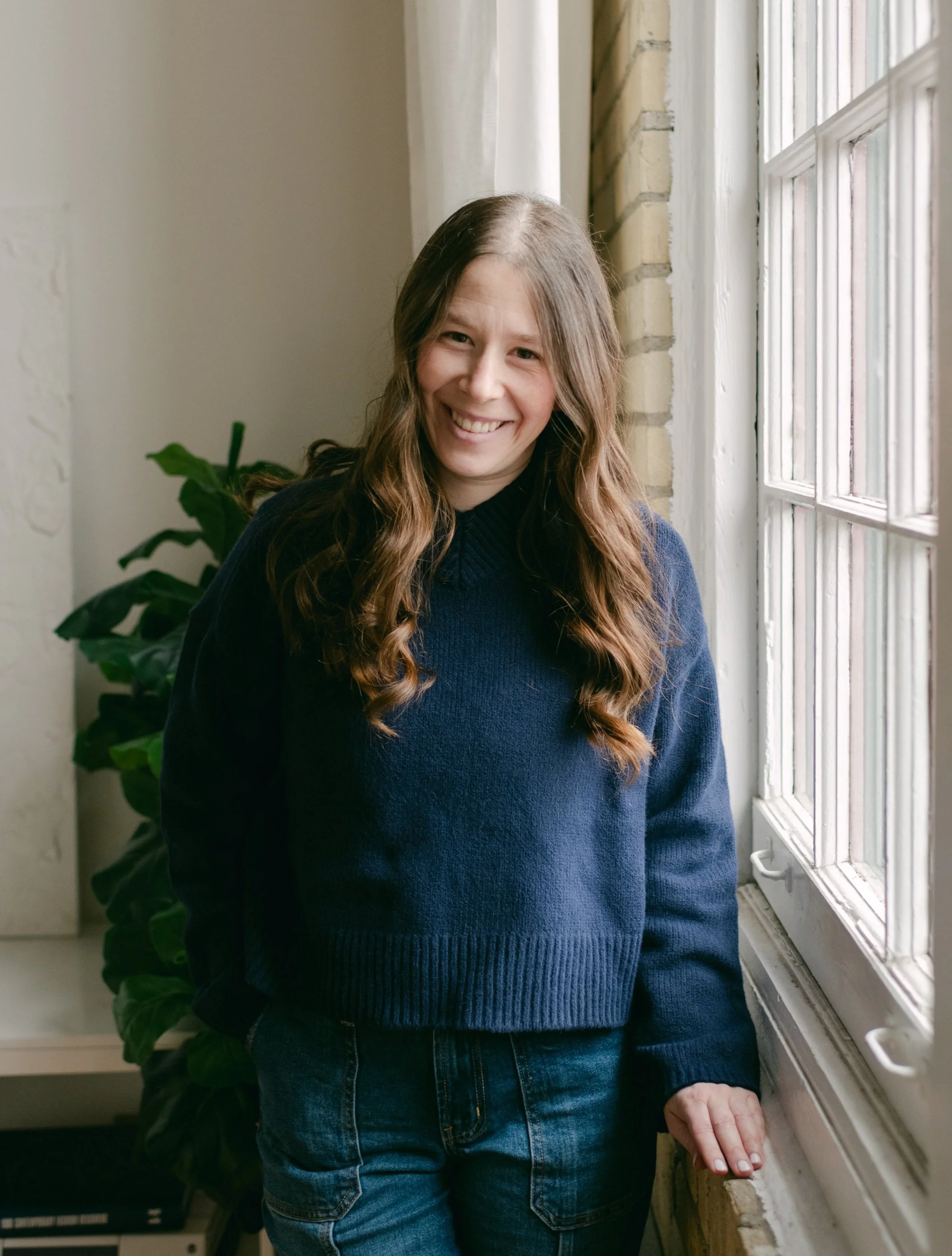 Young woman with long wavy brown hair smiling, wearing a navy sweater and jeans, standing by a window in a room with a plant behind her.