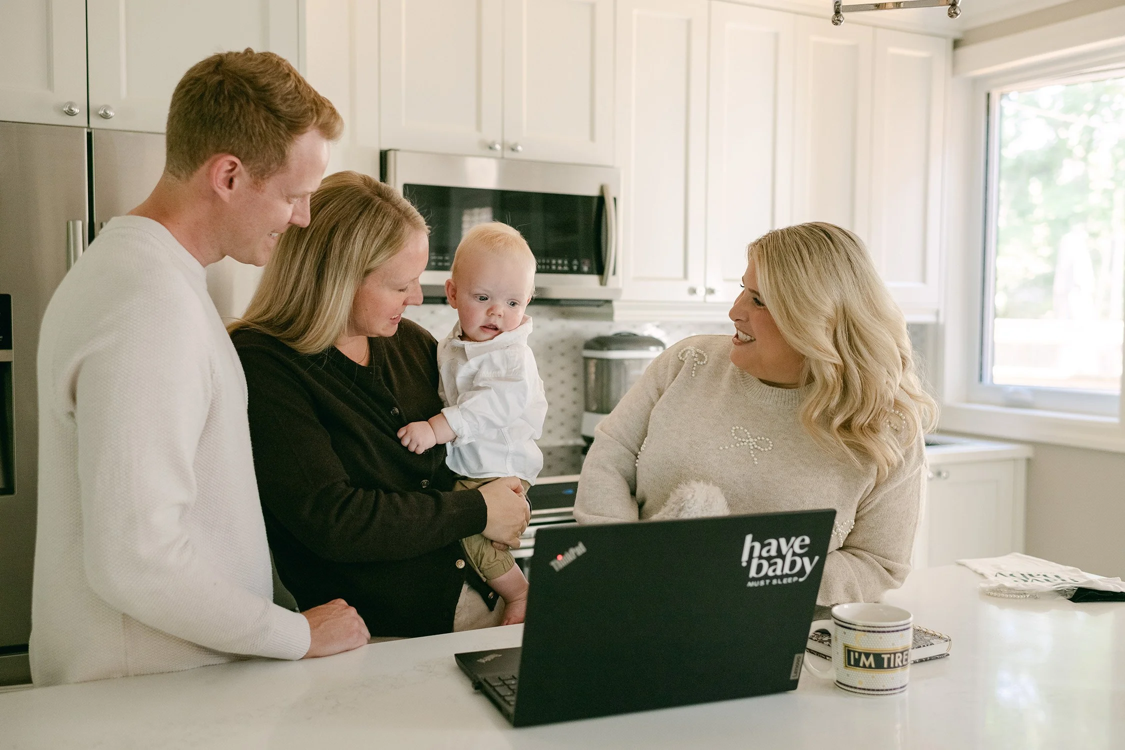 A family of four in a kitchen, including a woman holding a baby, a man, and another woman sitting at a table with a laptop and a mug, smiling and sharing a joyful moment.