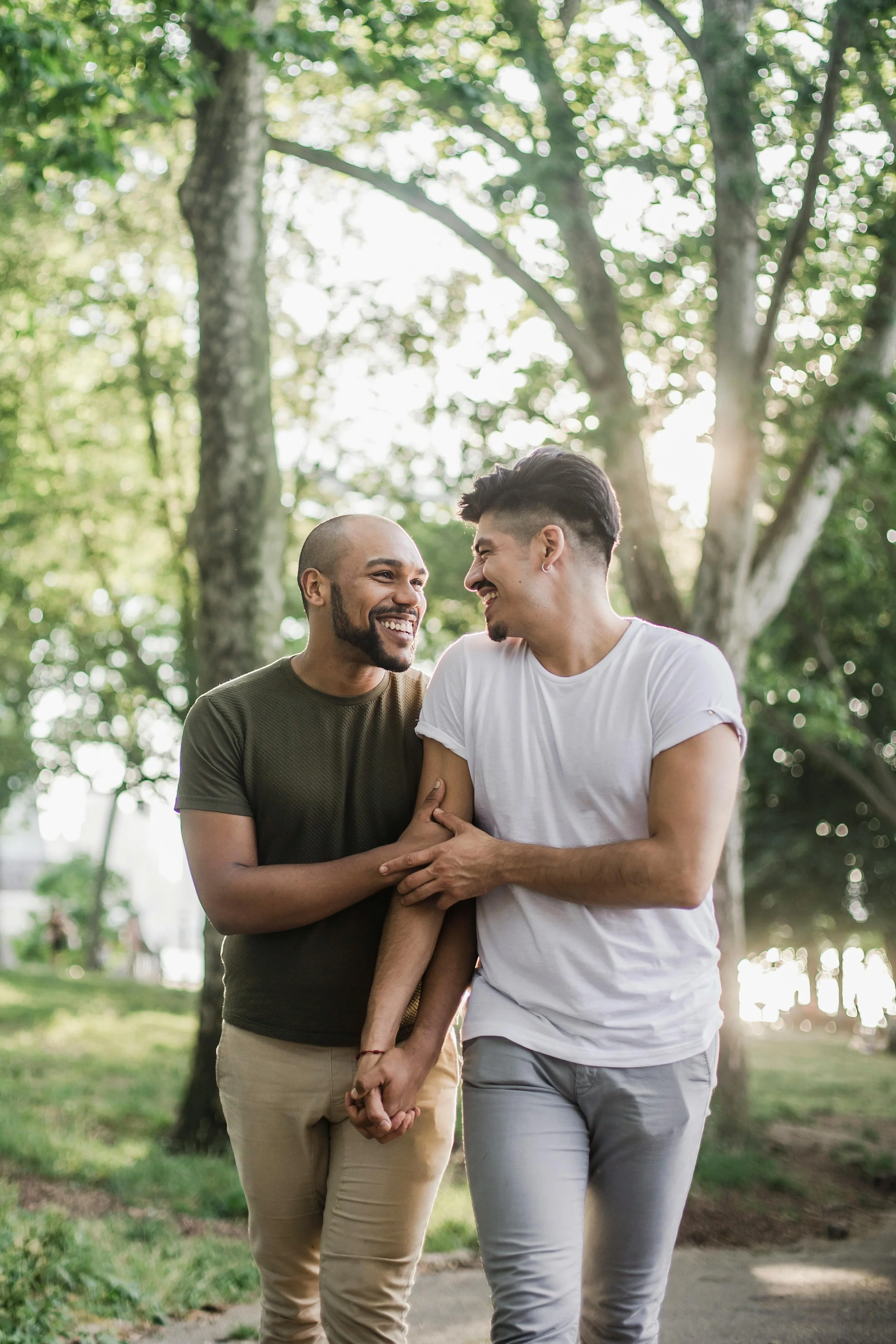 Two men are walking in a park, smiling and laughing while holding hands. One has a shaved head and beard, wearing a green T-shirt. The other has short dark hair and is wearing a white T-shirt. They are surrounded by green trees and sunlight filtering through the leaves.