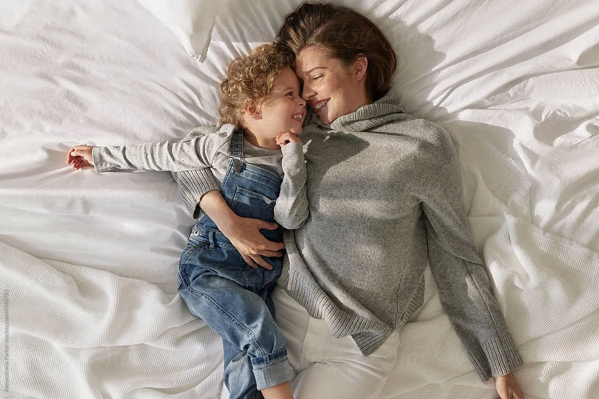 A woman and a young boy are lying on a bed, smiling and cuddling together. The woman is wearing a gray sweater and the boy is dressed in a light gray shirt with dark overalls and jeans.