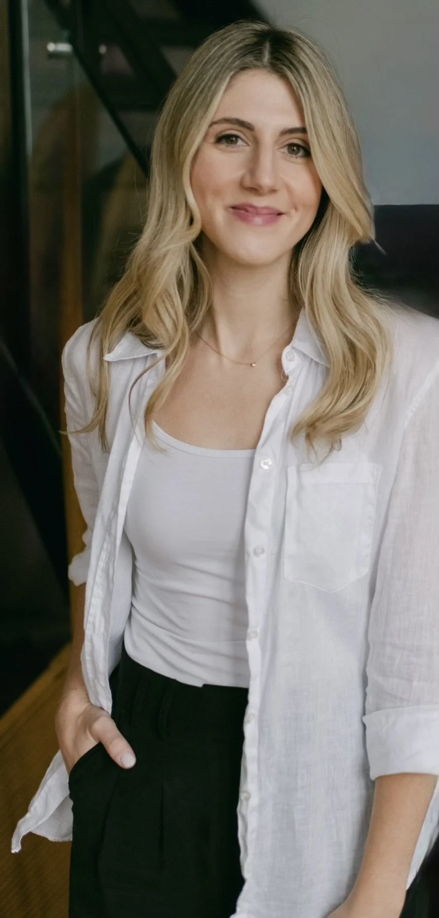 Woman with blonde hair wearing a white shirt and black pants, smiling and standing indoors.
