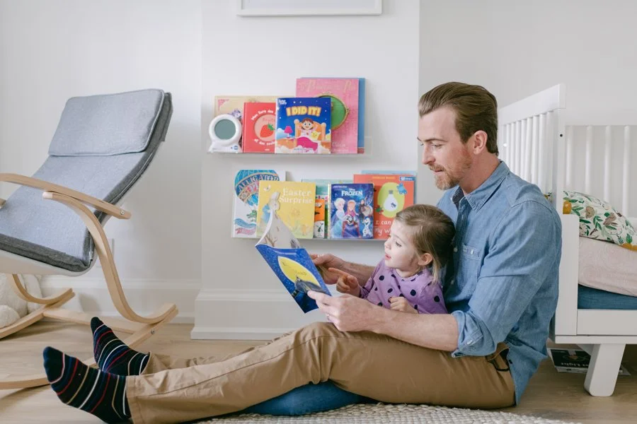 A man and young girl sitting on a rug in a child's bedroom, reading a book together. The girl is sitting on the man's lap and looking at the book. There are books and toys on a shelf behind them, and a rocking chair nearby.