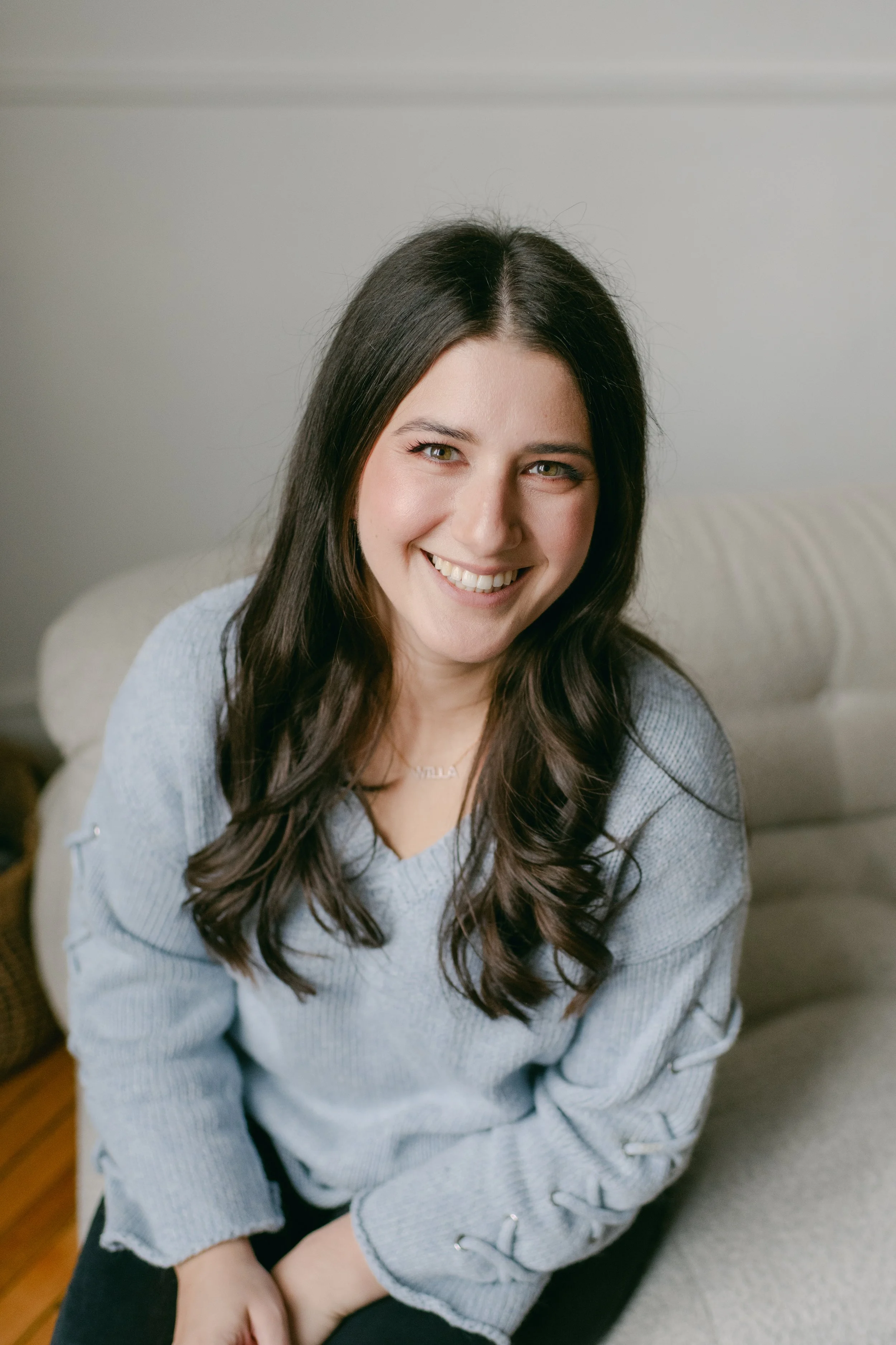 A woman with long, wavy dark brown hair smiling and looking at the camera, sitting on a light-colored couch in a cozy indoor setting.