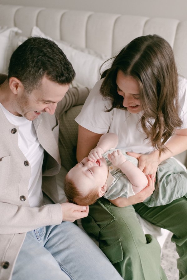 A couple sitting on a bed, smiling and playing with a laughing baby who is laying on the woman's lap.
