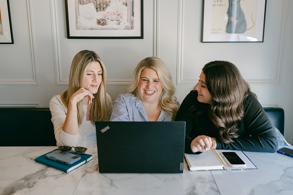 Three women sitting at a marble table, smiling and looking at a laptop screen. The table has notebooks, a phone, and sunglasses on it. They are in a room with framed pictures on the wall.