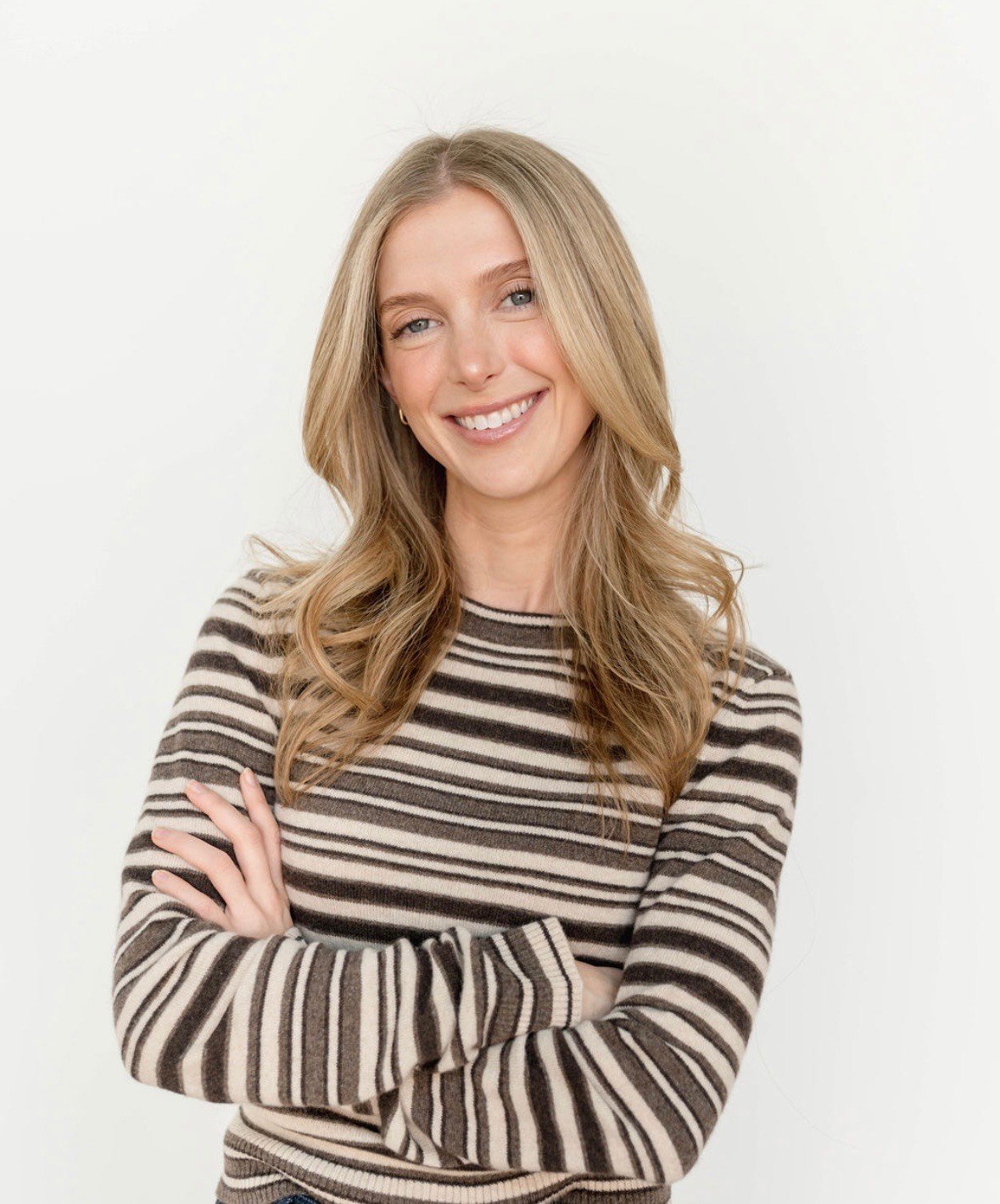 A smiling woman with long blonde hair wearing a striped beige and brown sweater, standing with arms crossed against a plain white background.