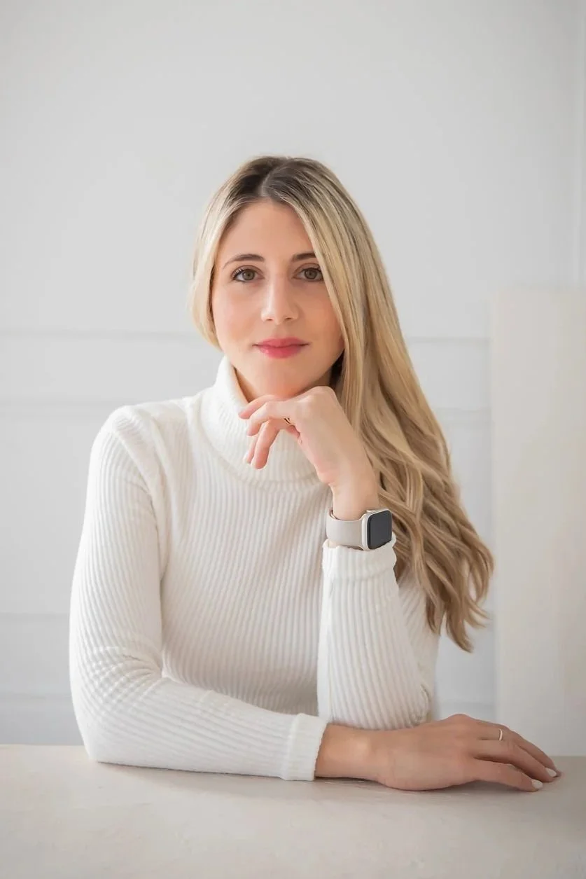 A woman with blonde hair wearing a white turtleneck sweater and a smartwatch, sitting at a table with her chin resting on her hand.