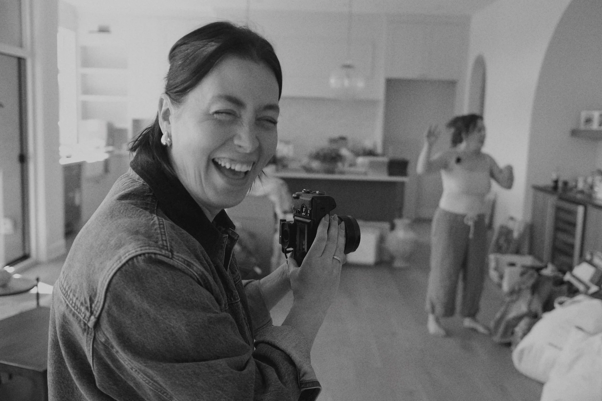 A woman laughing and holding a camera indoors, with a child in the background in a kitchen or dining area.