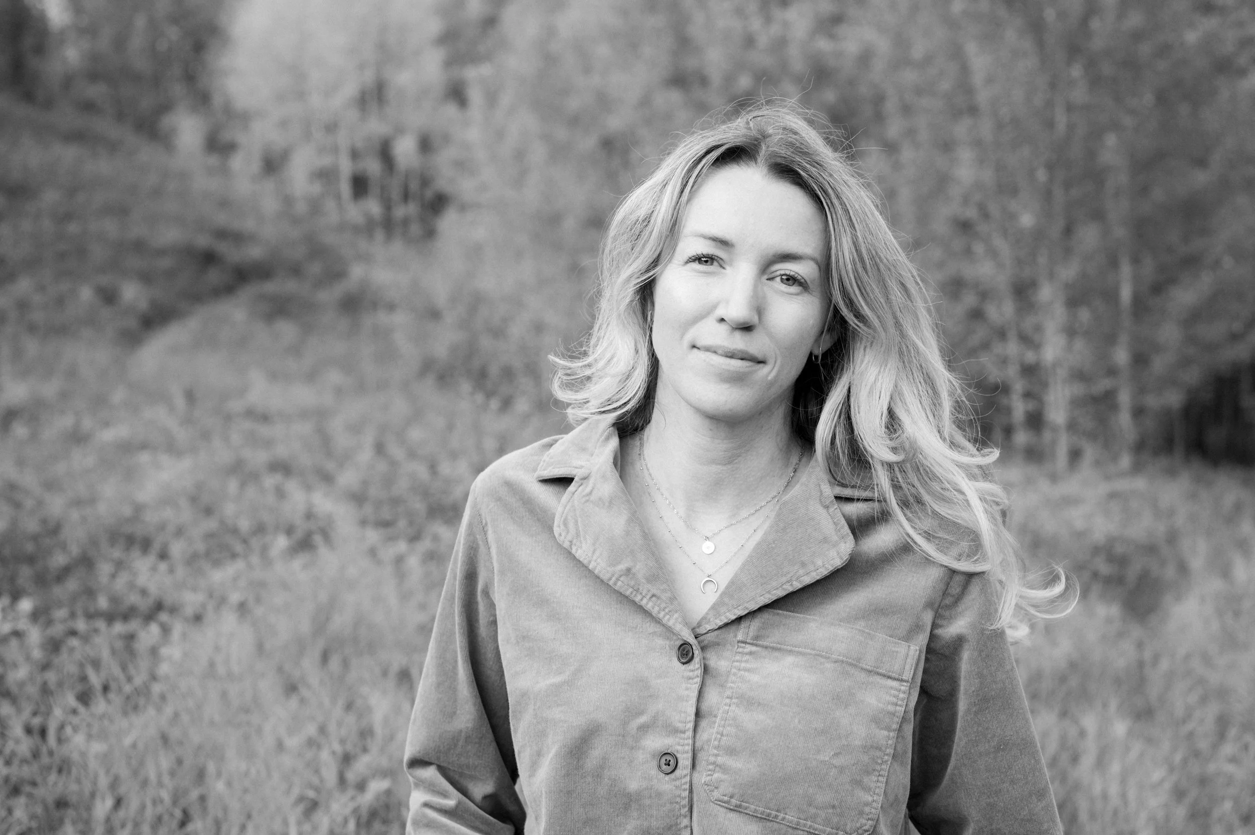 A woman with wavy hair standing outdoors in a grassy area with trees in the background, looking at the camera, in black and white.